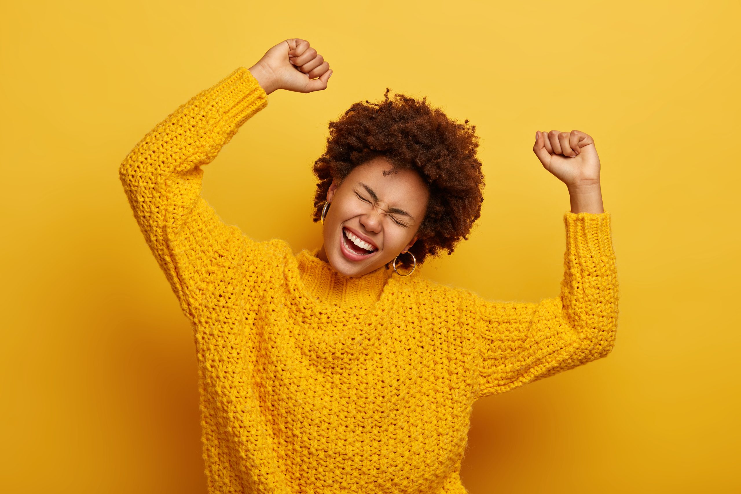 accomplishment-and-success-concept-joyful-afro-woman-raises-arms-feels-glad-and-relaxed-tilts-head-dressed-in-casual-knitted-jumper-laughs-from-happiness-celebrates-victory-isolated-on-yellow
